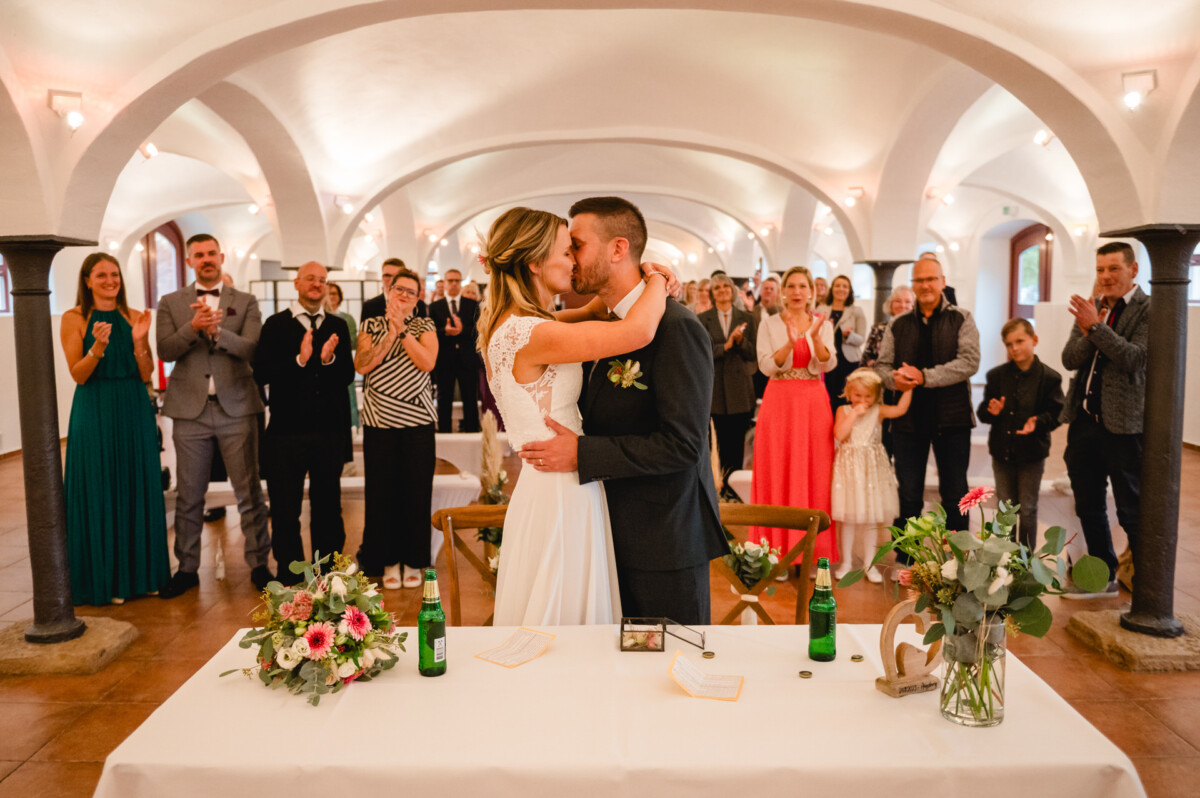 Braut und Bräutigam küssen sich in einem weißen, gewölbten Raum voller applaudierender Gäste. Auf dem Tisch im Vordergrund liegen ein Blumenarrangement und Papiere.