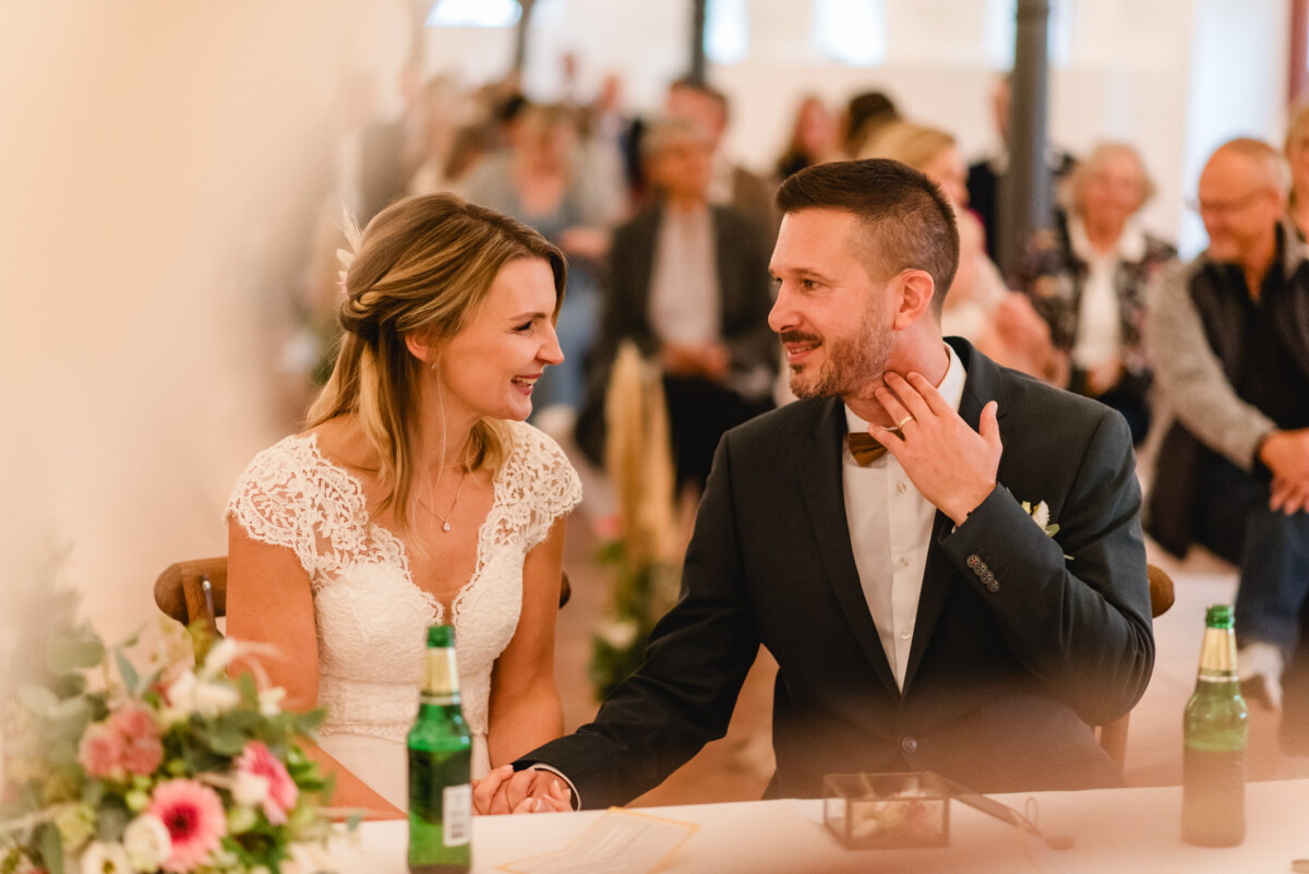 Braut und Bräutigam sitzen bei einer Hochzeitszeremonie Seite an Seite und halten Händchen. Die Braut trägt ein weißes Kleid und der Bräutigam einen Anzug mit Fliege. Sie sind von Gästen und Blumendekorationen umgeben.