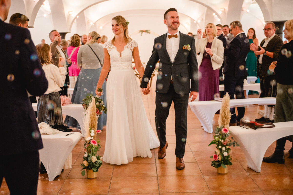 Braut und Bräutigam gehen in einem dekorierten Innenraum Händchen haltend den Gang entlang, während die Gäste applaudieren. Seifenblasen schweben in der Luft.