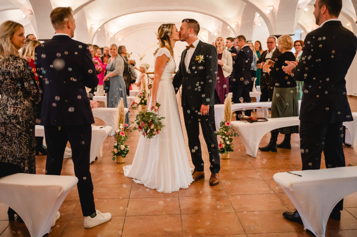 Braut und Bräutigam küssen sich in einem dekorierten Saal, umgeben von stehenden Gästen.