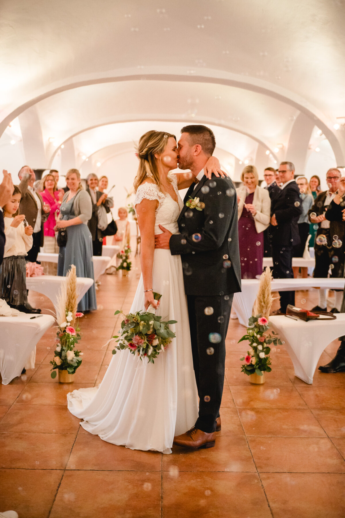 Braut und Bräutigam küssen sich bei ihrer Hochzeitszeremonie, umgeben von Gästen in einem dekorierten Saal mit herumschwebenden Seifenblasen.