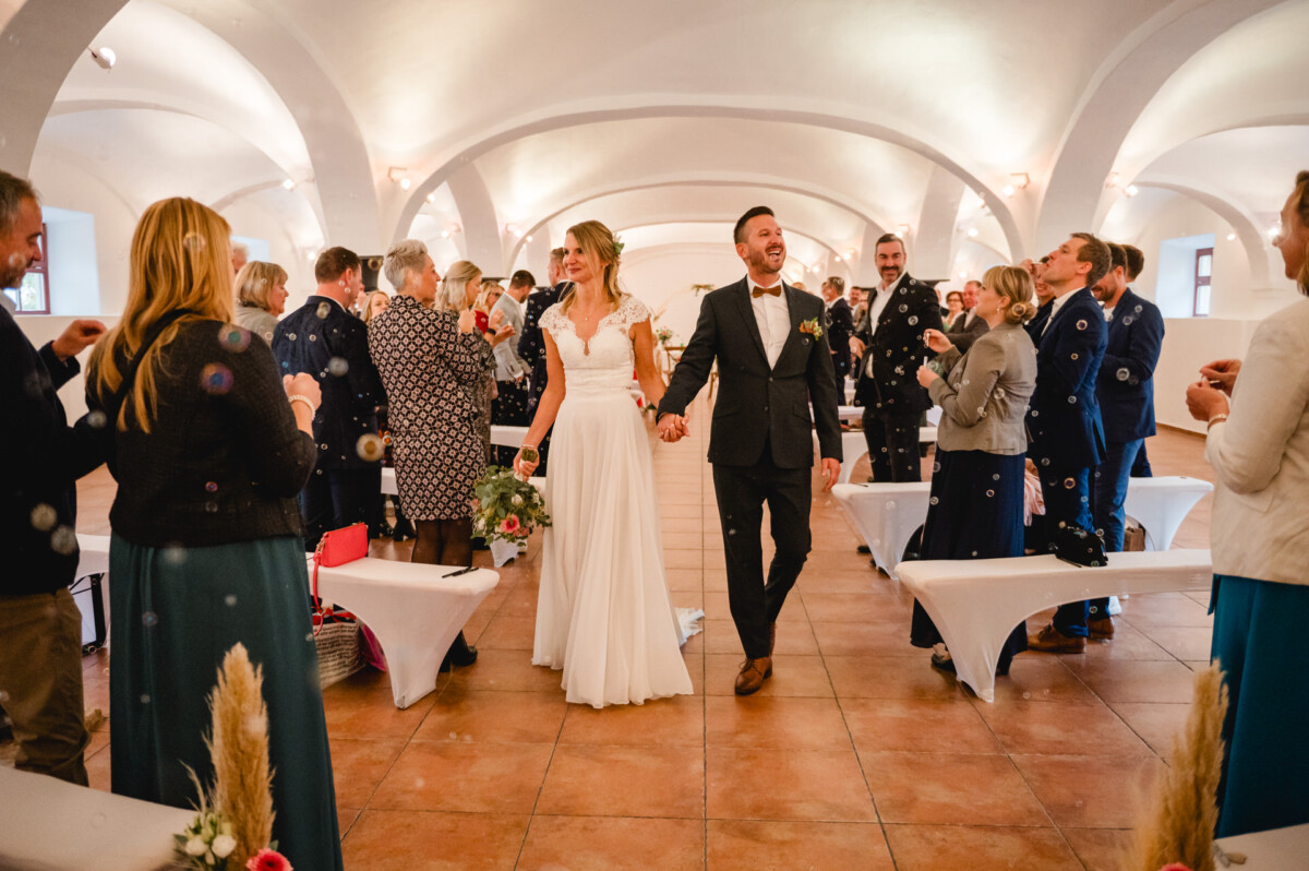 Eine Braut und ein Bräutigam schreiten lächelnd in einem geschmückten Saal den Gang entlang. Gäste stehen auf beiden Seiten, klatschen und machen Fotos.