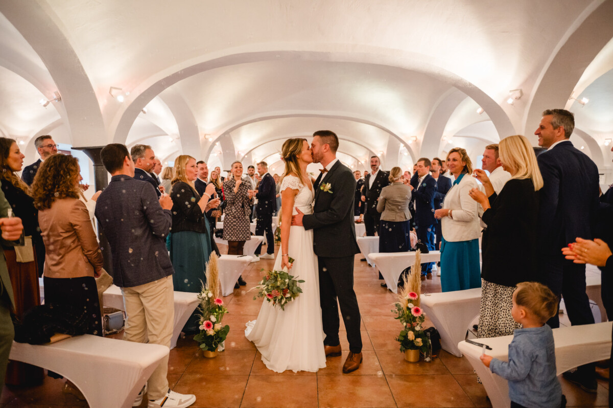 Ein Paar küsst sich am Ende einer Hochzeitszeremonie in einem Gewölberaum, umgeben von applaudierenden Gästen.