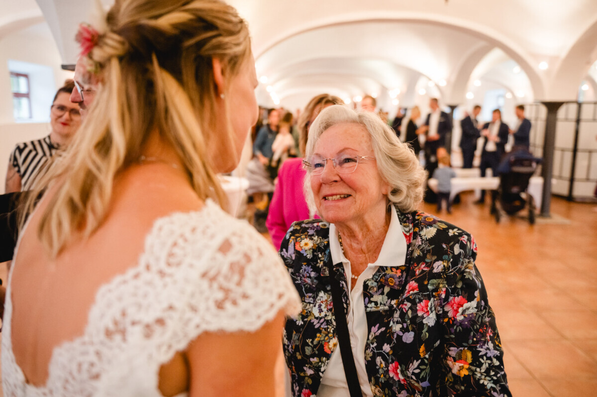 Eine lächelnde ältere Frau spricht bei einer Zusammenkunft in einem dekorierten Saal mit einer Braut im Spitzenkleid. Im Hintergrund sind Menschen zu sehen.
