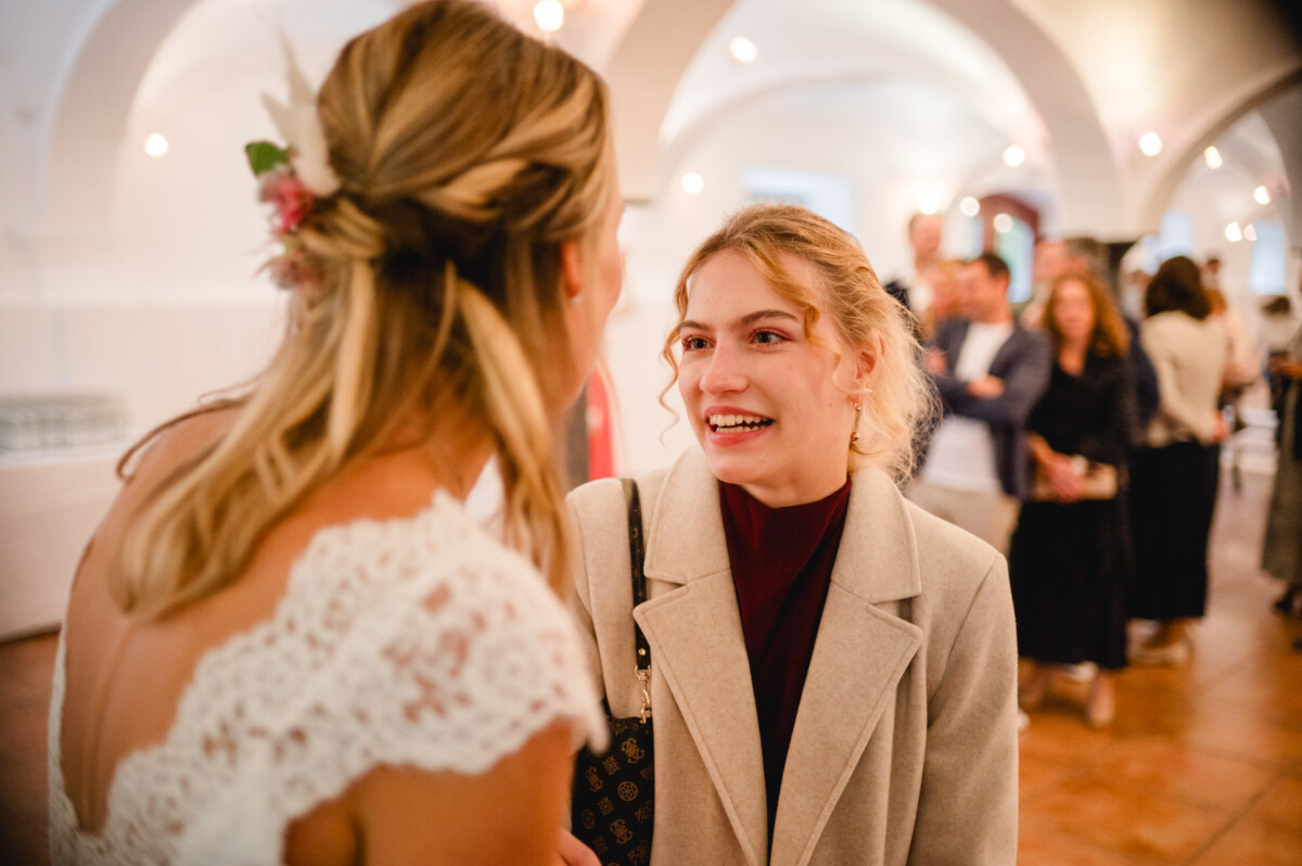 Zwei Frauen lächeln und unterhalten sich bei einer Versammlung in geschlossenen Räumen, im Hintergrund sind die Menschen unscharf.
