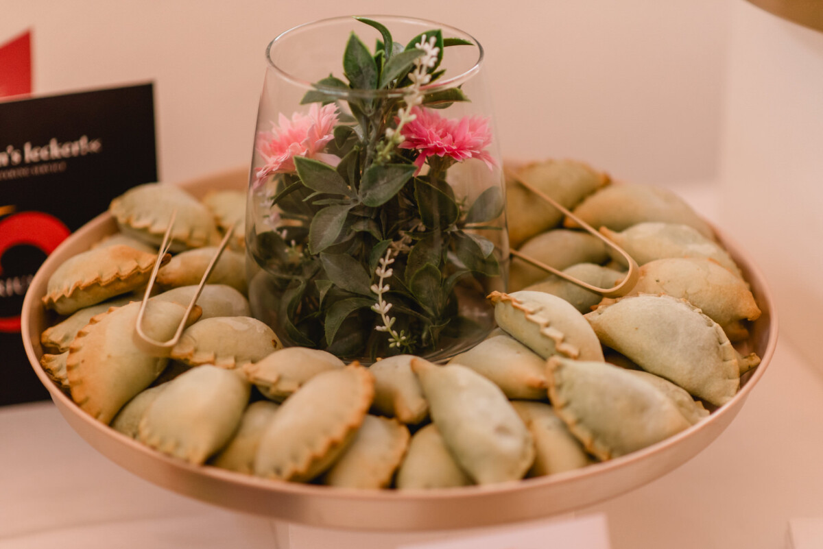 Ein rundes Tablett mit grünen Empanadas, die eine Glasvase mit rosa Blumen und Grünpflanzen umgeben.