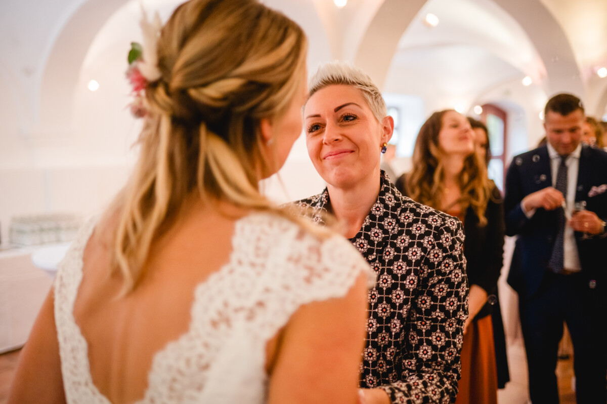 Bei einer Indoor-Veranstaltung stehen sich zwei Personen gegenüber. Eine trägt ein Spitzenkleid und die andere eine gemusterte Jacke. Die Personen im Hintergrund sind in Abendgarderobe gekleidet.