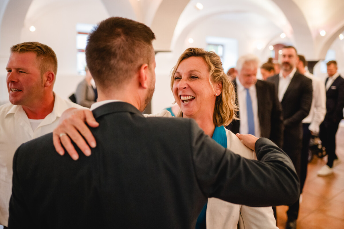 Eine Frau und ein Mann in Abendgarderobe umarmen sich fröhlich bei einem geselligen Beisammensein, während sich im Hintergrund andere Leute unterhalten.