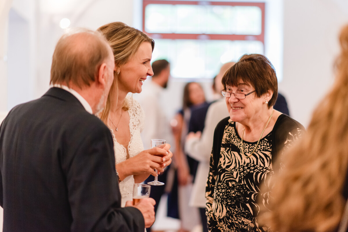 Drei Personen unterhalten sich bei einer gesellschaftlichen Veranstaltung. Eine Frau hält ein Glas und alle scheinen zu lächeln. Im unscharfen Hintergrund sind Gäste zu sehen.