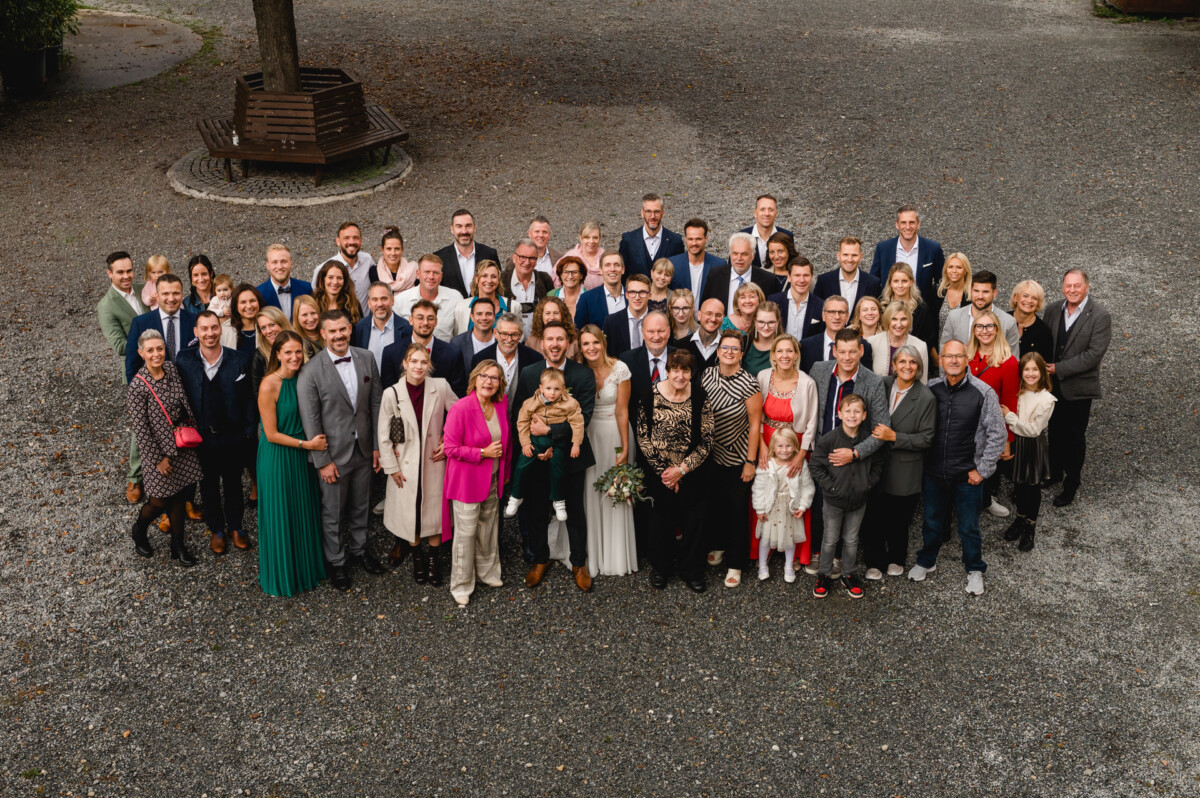 Eine große Gruppe von Menschen posierte gemeinsam im Freien auf einer Kiesfläche, mit Bäumen im Hintergrund.