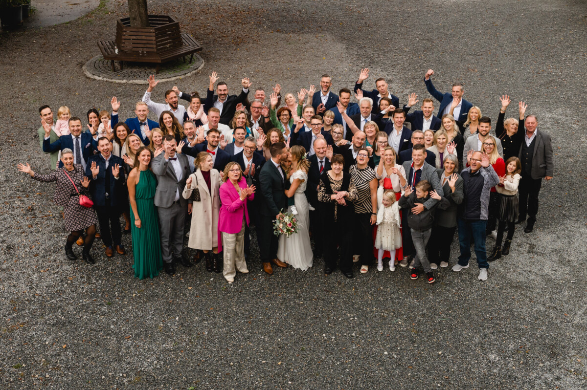 Eine große Gruppe von Menschen, darunter ein Paar im Hochzeitskleid, steht auf einer Kiesfläche, lächelt und winkt in die Kamera.