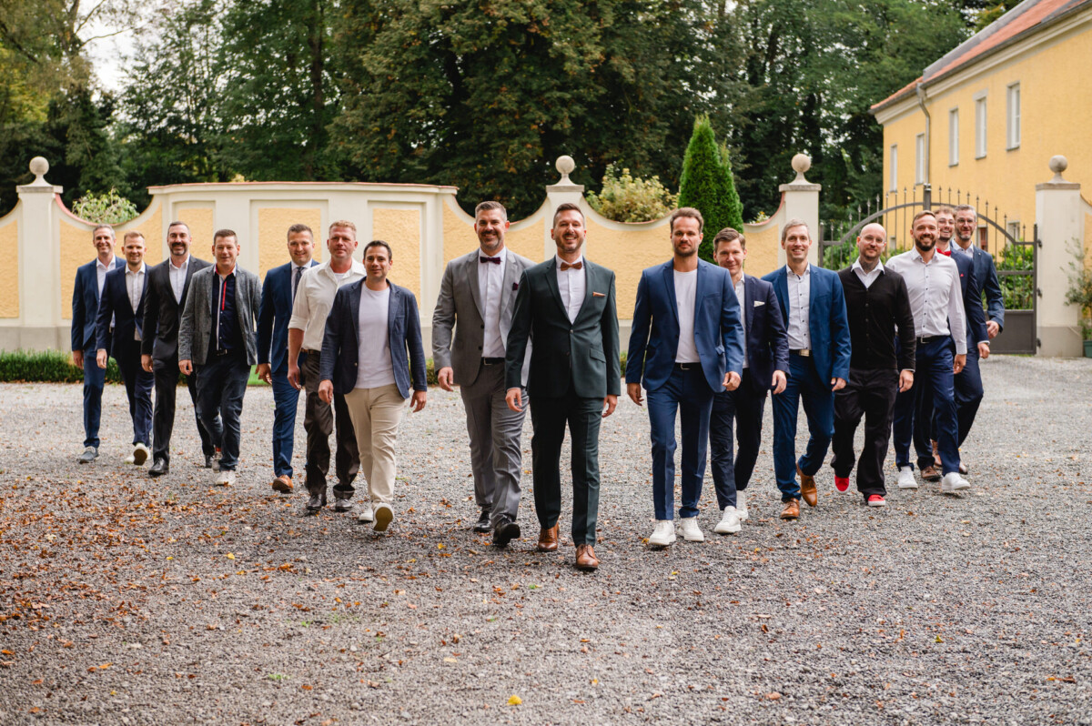 Eine Gruppe von Männern in legerer Geschäftskleidung geht gemeinsam im Freien auf einem Kiesweg spazieren, umgeben von grünen Bäumen und gelben Gebäuden.