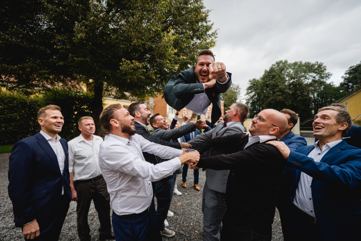 Eine Gruppe von Männern in Anzügen hebt draußen einen anderen Mann in die Luft und wirkt dabei feierlich. Im Hintergrund sind Bäume und Gebäude zu sehen.