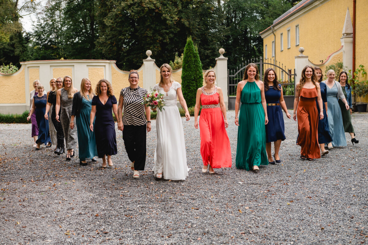 Eine Braut und ihre Hochzeitsgesellschaft gehen im Freien auf einem Kiesweg in der Nähe gelber Gebäude spazieren. Sie tragen bunte Kleider und halten einen Blumenstrauß in den Händen.