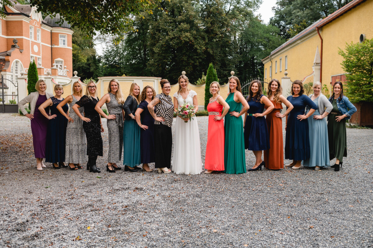 Eine Braut in Weiß steht mit 13 Frauen, alle in unterschiedlich bunten Kleidern, im Freien auf einem Kiesweg mit Bäumen und Gebäuden im Hintergrund.