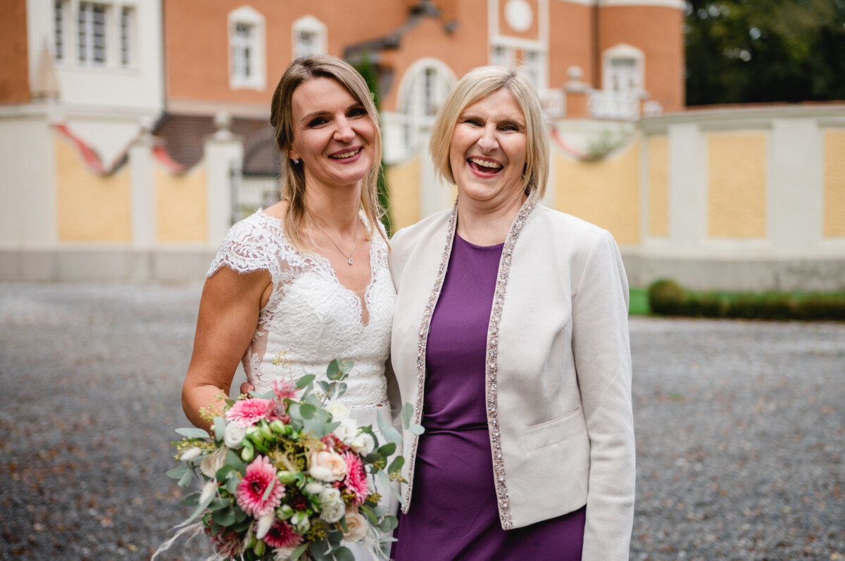 Zwei Frauen stehen im Freien und lächeln. Eine trägt ein weißes Spitzenkleid und hält einen Blumenstrauß; die andere trägt ein lila Kleid und eine leichte Jacke. Im Hintergrund ist ein Gebäude mit orangefarbenen und gelben Wänden zu sehen.