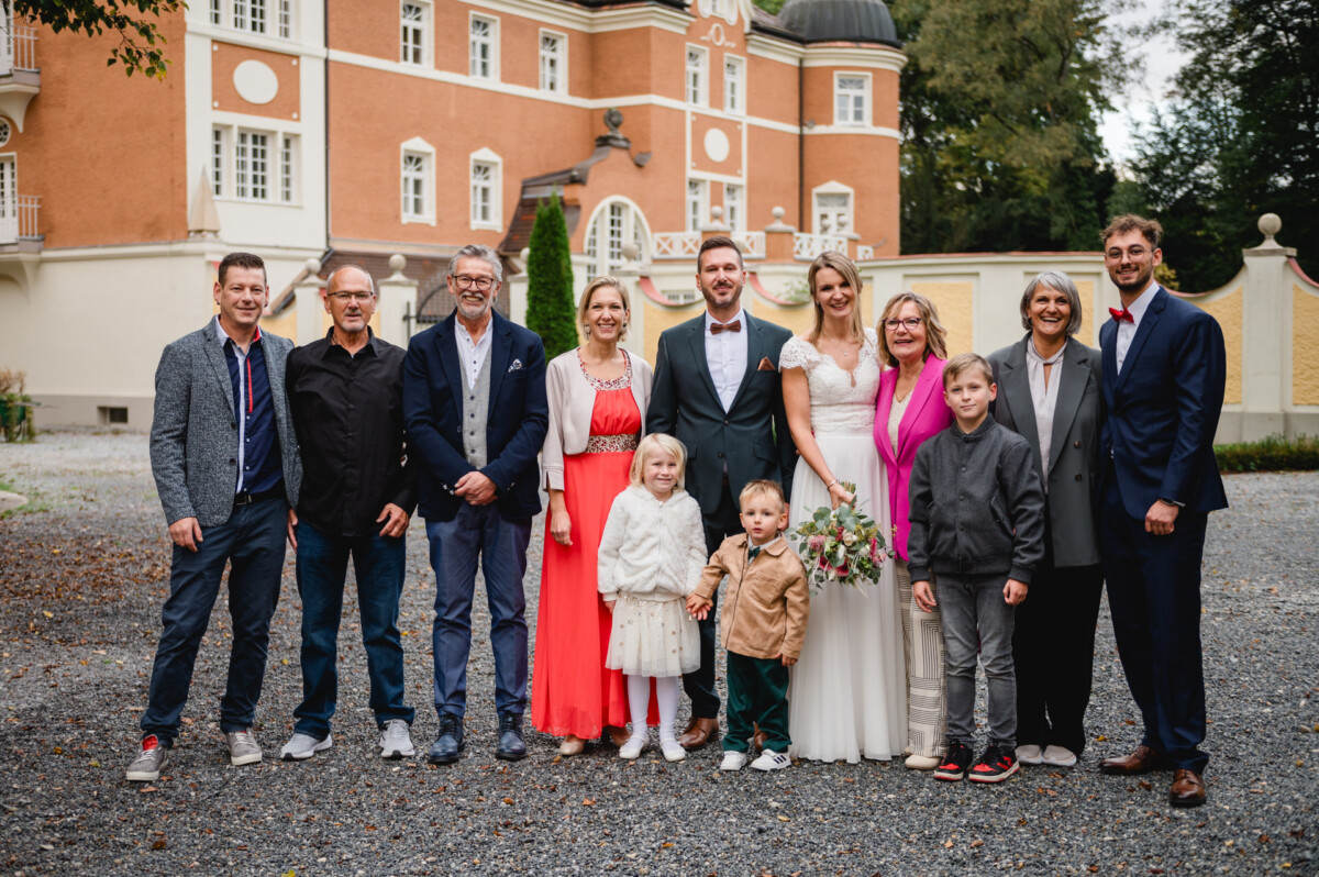 Eine Gruppe von Menschen, darunter Braut und Bräutigam, steht für ein Foto gemeinsam im Freien vor einem historischen Gebäude.