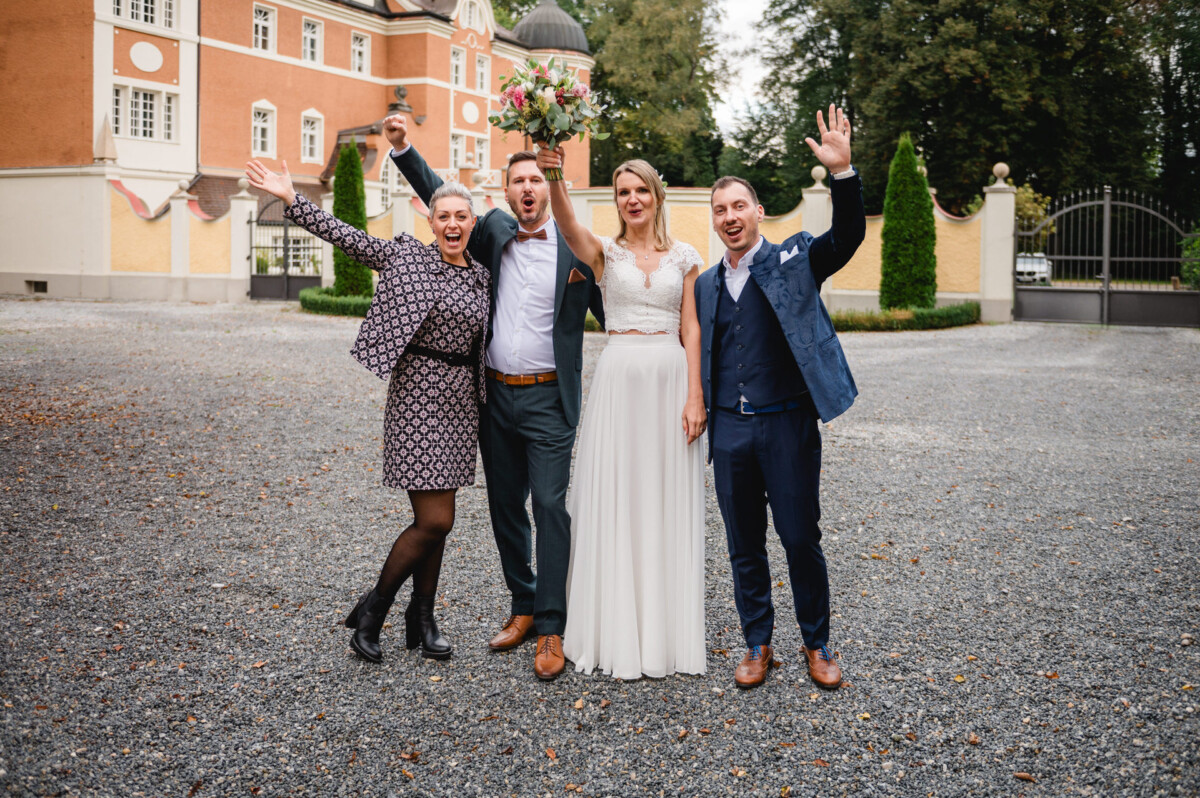 Vier Menschen, darunter Braut und Bräutigam, stehen gemeinsam auf einem Kiesweg vor einem großen Gebäude, alle lächeln und heben feierlich die Arme.