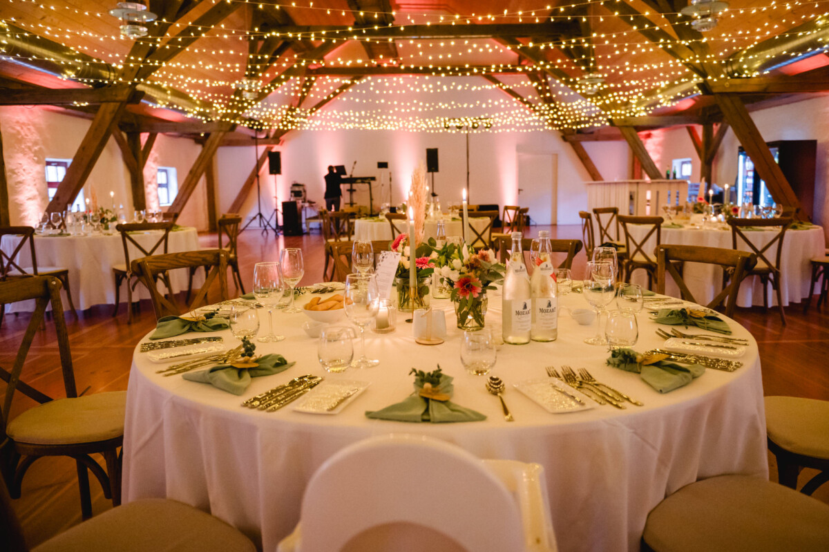 Ein dekorierter Veranstaltungsraum mit runden Tischen zum Essen, Blumengestecken und sanfter Beleuchtung durch Lichterketten an Holzbalken.