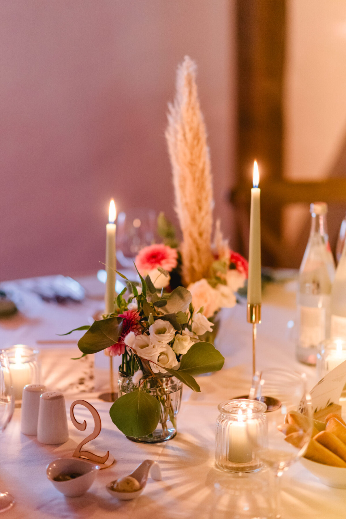 Elegante Tischdekoration mit brennenden Kerzen, Blumenschmuck, Besteck und Wasserflaschen auf einer weißen Tischdecke.