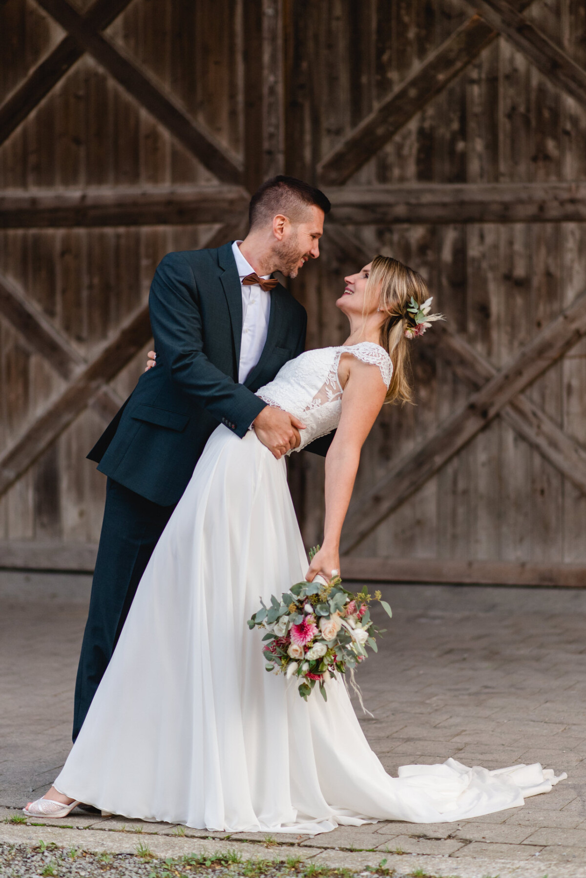 Ein Bräutigam im dunklen Anzug taucht eine Braut in einem weißen Kleid mit Blumenkopfschmuck und Blumenstrauß vor einer rustikalen Holztür ins Wasser.