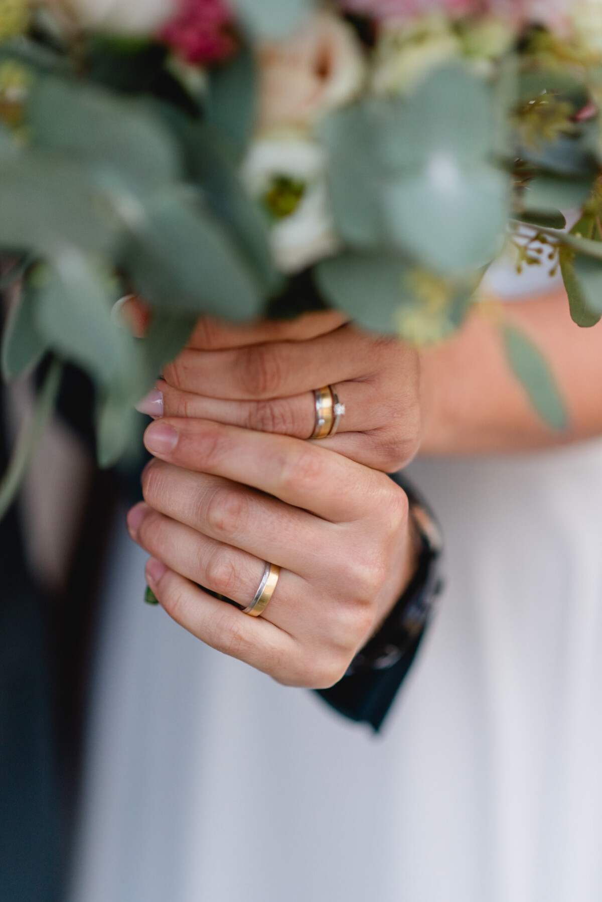 Nahaufnahme von zwei Personen, die einen Blumenstrauß mit sichtbaren Eheringen an ihren Fingern halten.