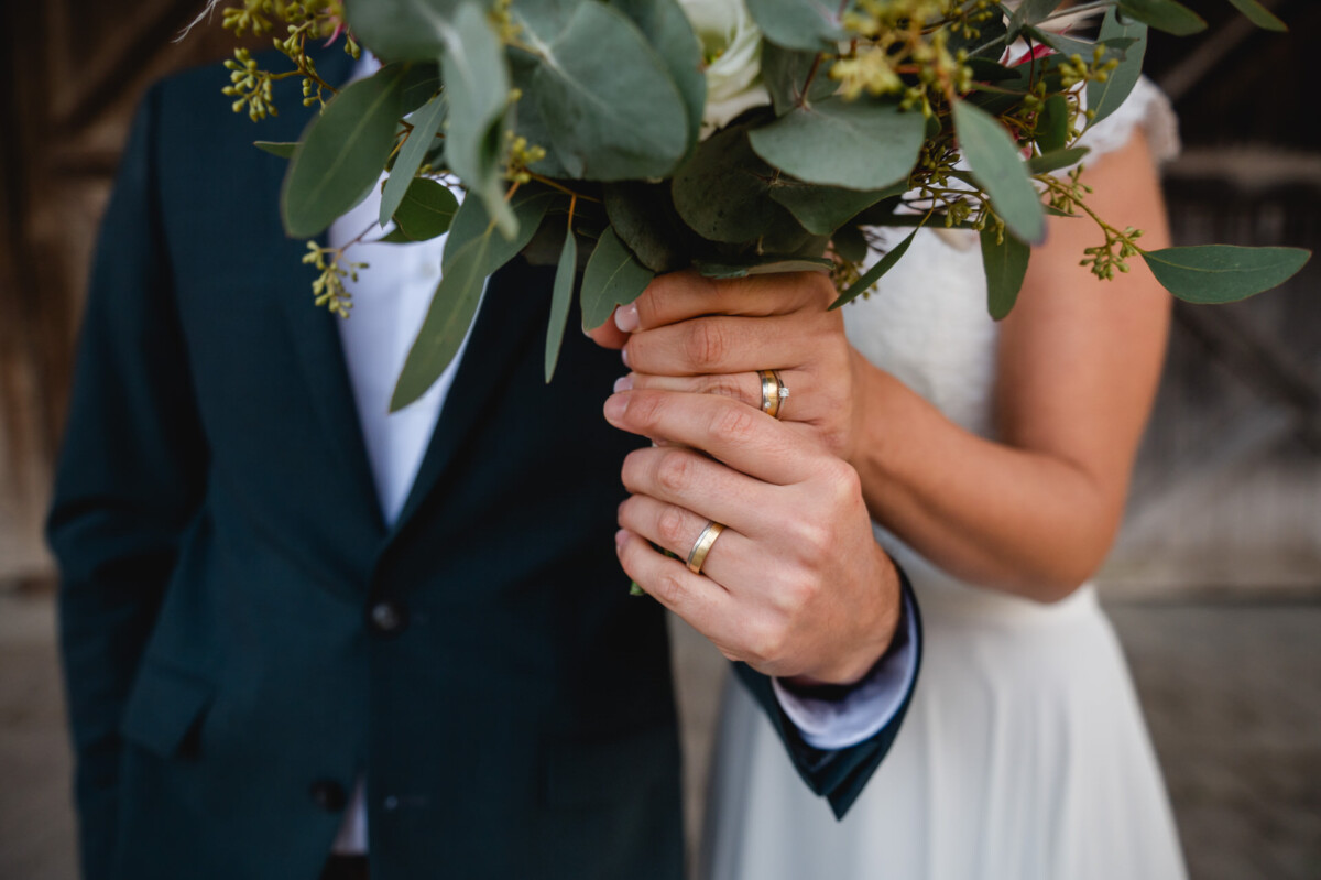 Ein Paar im Hochzeitskleid, beide zeigen ihre Eheringe. Sie halten einen großen Strauß aus grünen Blättern und weißen Blumen.