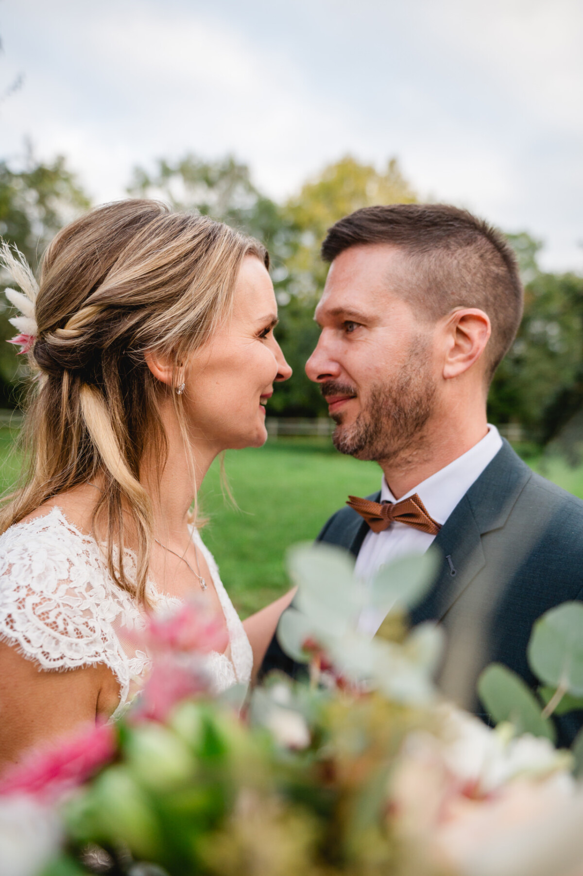 Ein Paar in Hochzeitskleidung steht dicht beieinander, vor einem grasbewachsenen Hintergrund. Die Frau trägt ein Spitzenkleid und der Mann einen Anzug mit Fliege. Im Vordergrund sind Blumen zu sehen.