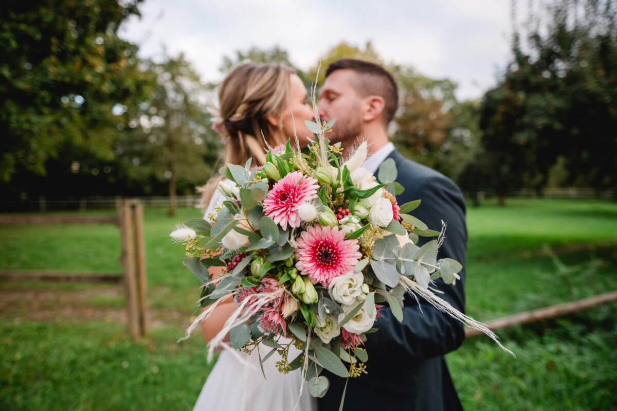 Ein Paar küsst sich im Freien und hält einen Strauß rosafarbener und weißer Blumen in der Hand. Im Vordergrund sieht man Grünpflanzen. Im Hintergrund sind Gras, Bäume und ein eingezäunter Bereich zu sehen.
