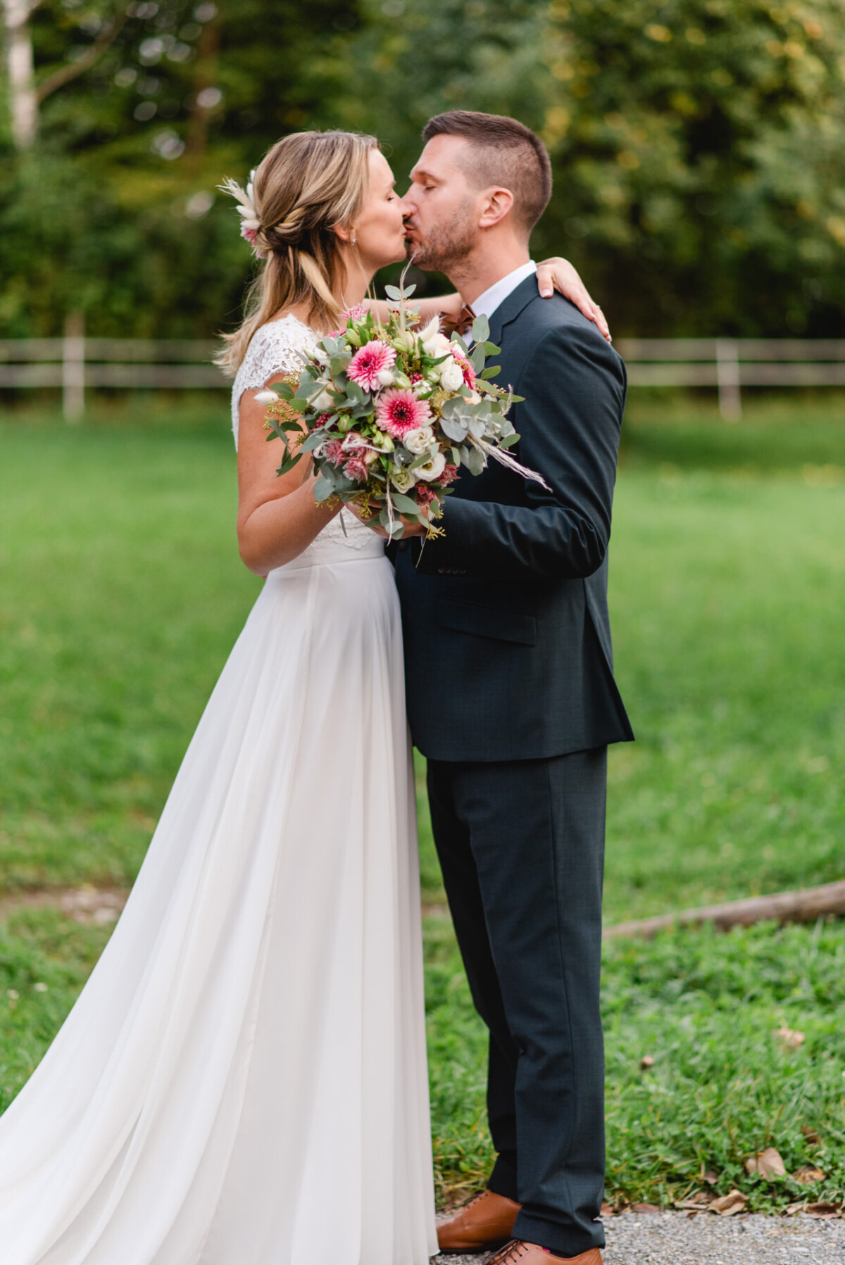 Braut und Bräutigam küssen sich in einem Garten, die Braut hält einen Strauß rosa und weißer Blumen, beide in Abendgarderobe.
