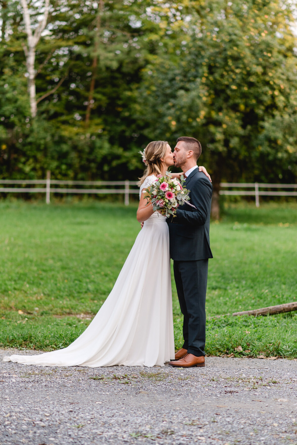 Braut und Bräutigam küssen sich im Freien auf einem Kiesweg. Die Braut hält einen Blumenstrauß in der Hand. Im Hintergrund sind Bäume und Gras zu sehen.