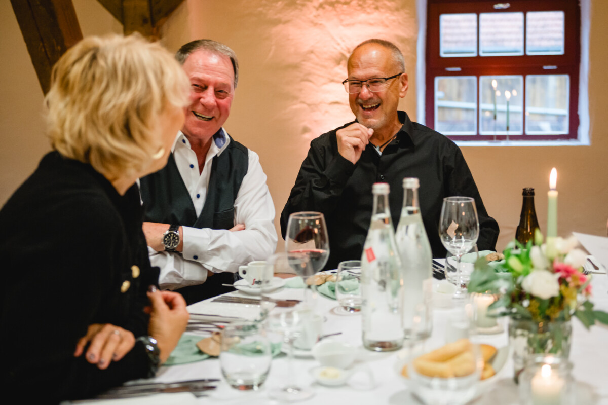 Drei Personen sitzen lachend und unterhalten sich an einem Esstisch. Der Tisch ist mit Kerzen, Gläsern und Wasserflaschen gedeckt.