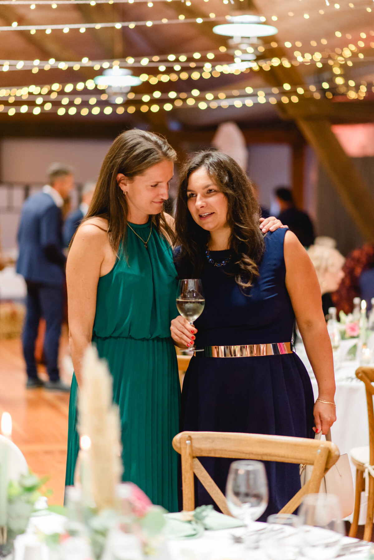 Zwei Frauen in Kleidern stehen bei einer dekorierten Veranstaltung im Innenbereich zusammen. Eine hält ein Weinglas. Darüber hängen Lichterketten und im Hintergrund sind Tische aufgestellt.