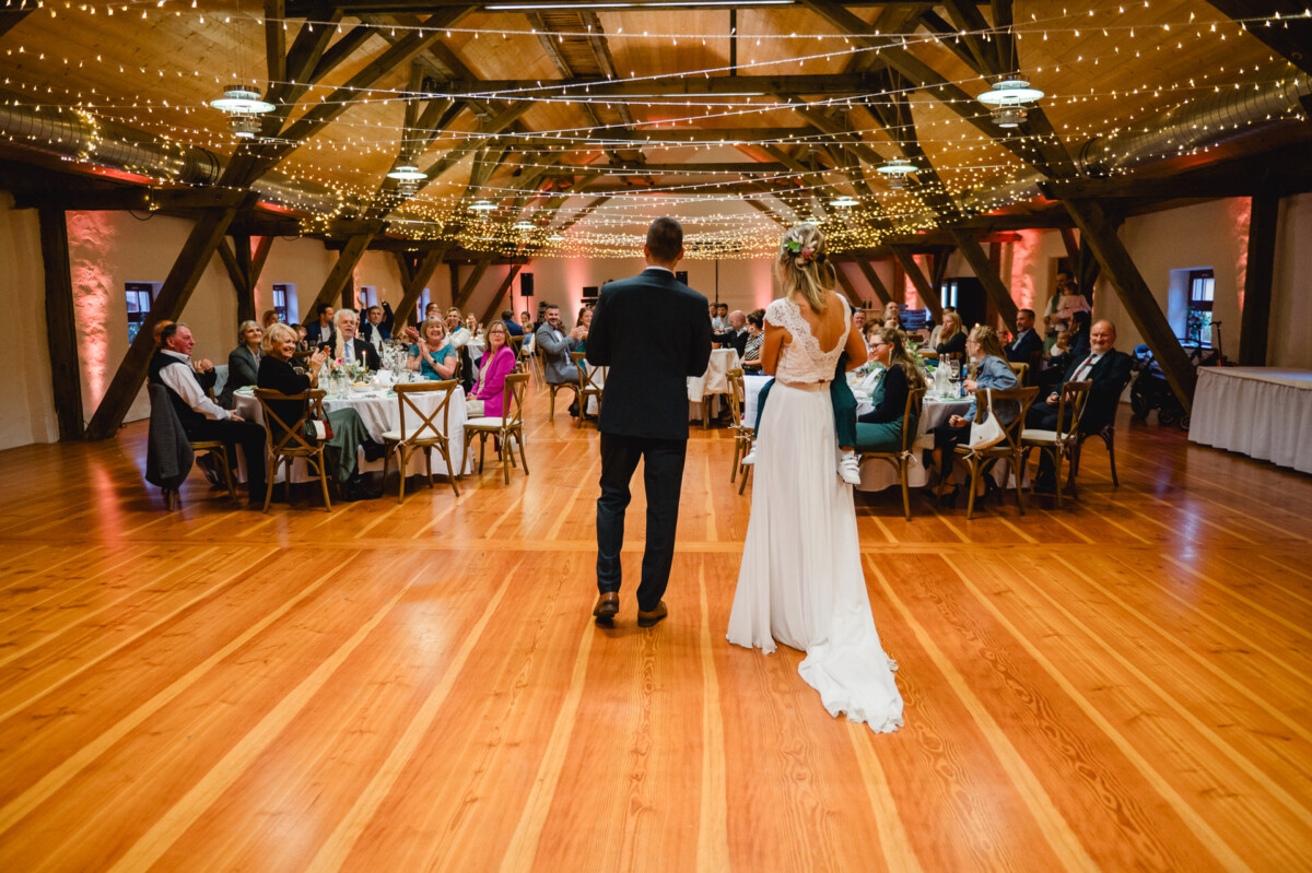 Eine Braut und ein Bräutigam stehen in einer funkelnden, dekorierten Halle und sprechen zu den sitzenden Gästen bei einem Hochzeitsempfang.