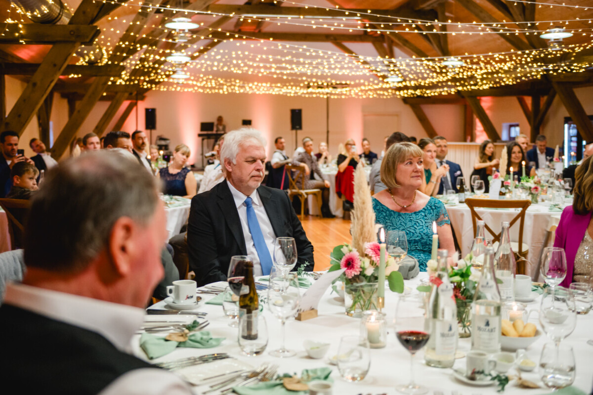Menschen sitzen an dekorierten Tischen in einem warm beleuchteten Veranstaltungsort mit Lichterketten darüber und nehmen an einer offiziellen Veranstaltung teil.