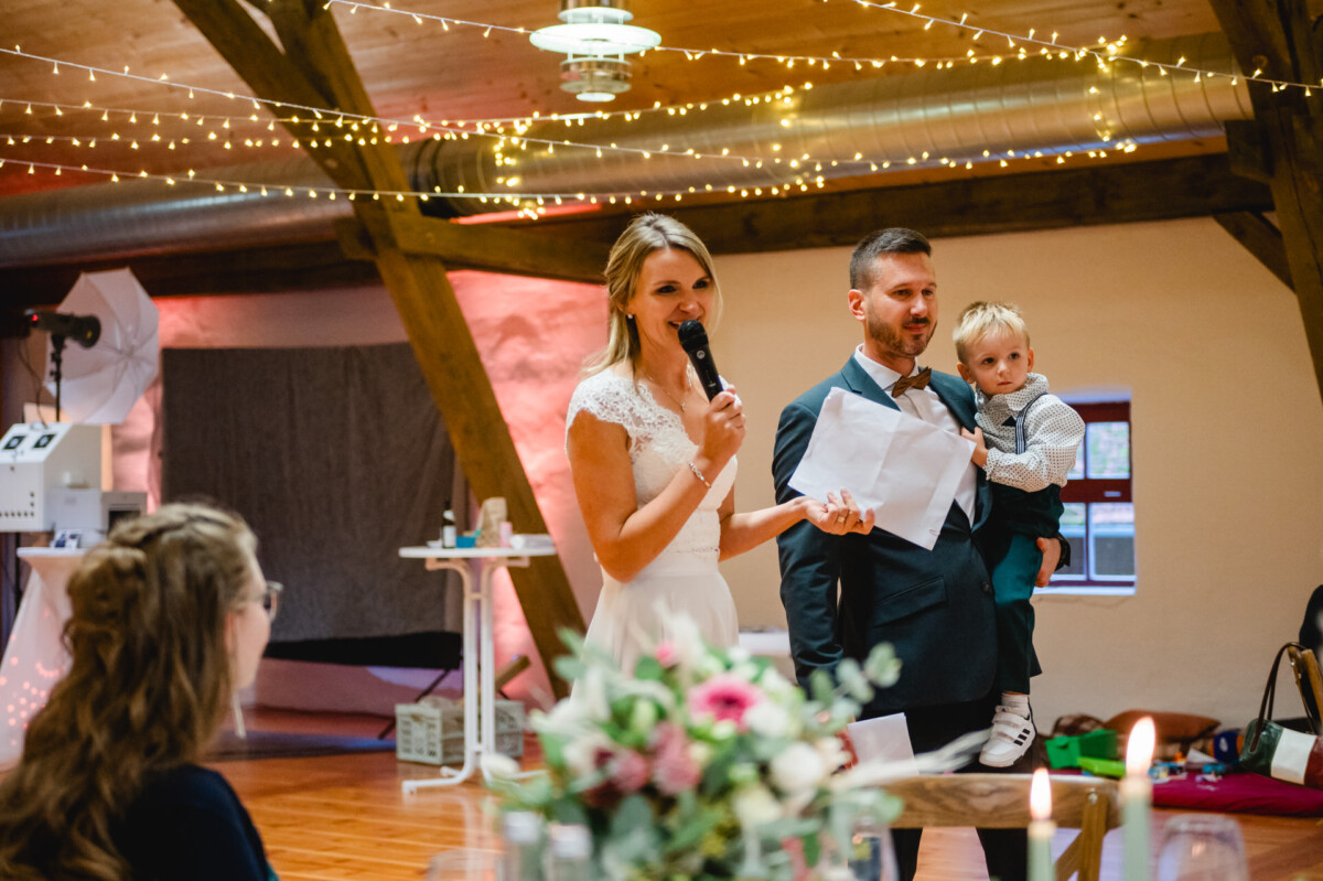 Eine Frau in einem weißen Kleid spricht in ein Mikrofon, während ein Mann im Anzug in einem dekorierten Veranstaltungsort ein Kind hält.