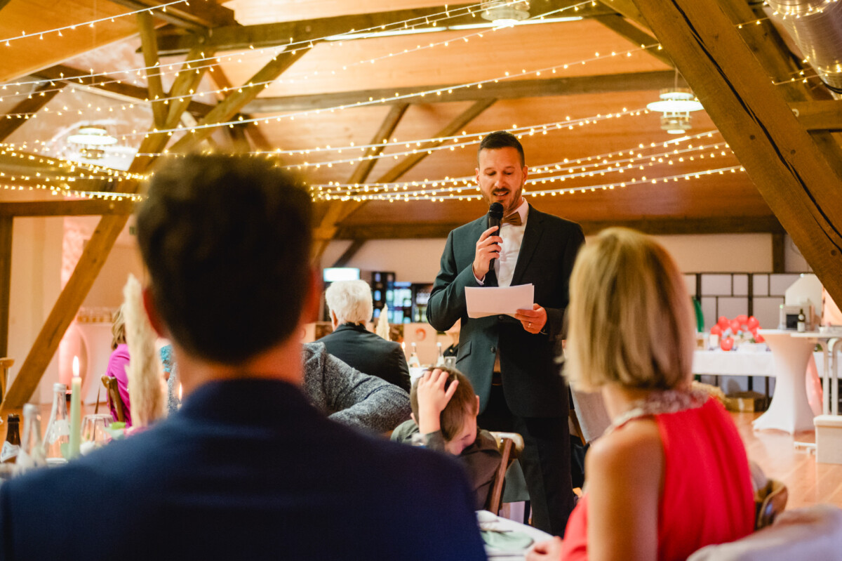 Ein Mann im Anzug spricht in ein Mikrofon und hält dabei ein Blatt Papier in der Hand. Er steht in einem warm beleuchteten Raum, der mit Lichterketten geschmückt ist. Die Leute sitzen an Tischen und hören ihm zu.
