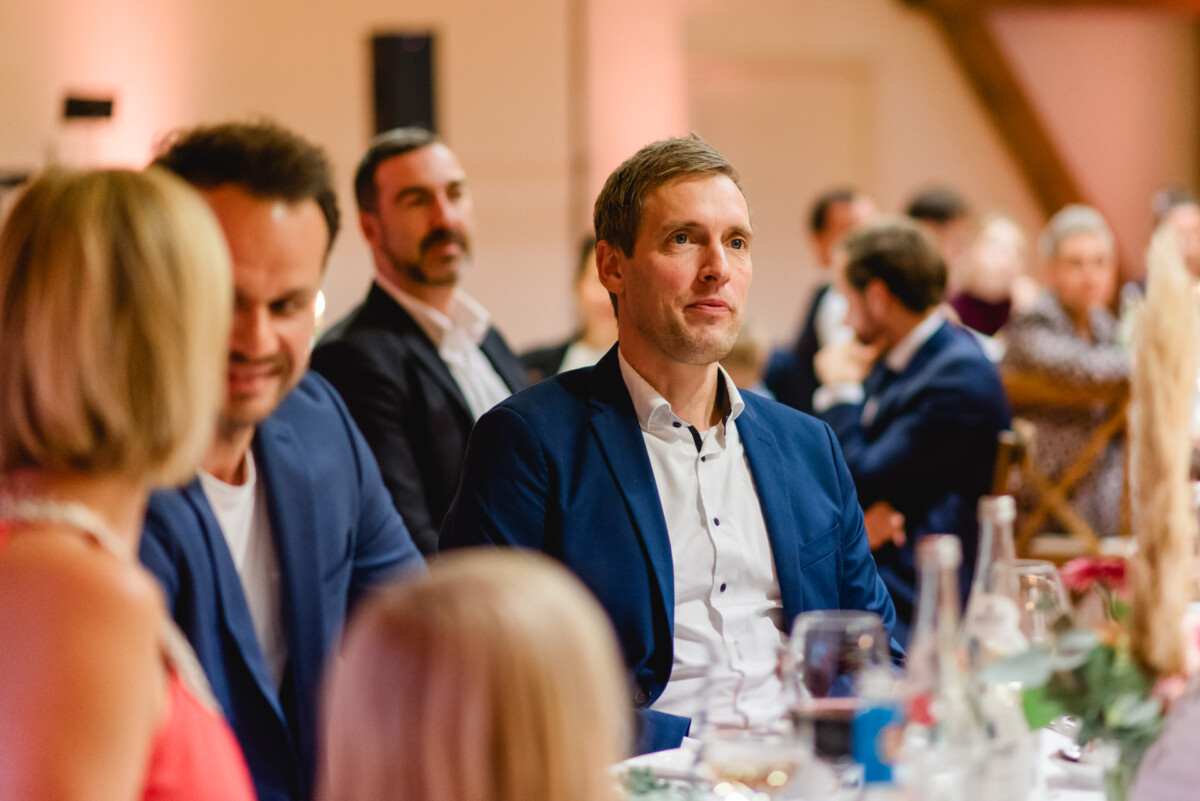 Eine Gruppe von Menschen in Abendgarderobe sitzt mit Gläsern und Flaschen an einem Tisch und hört in einem warm beleuchteten Raum aufmerksam zu.
