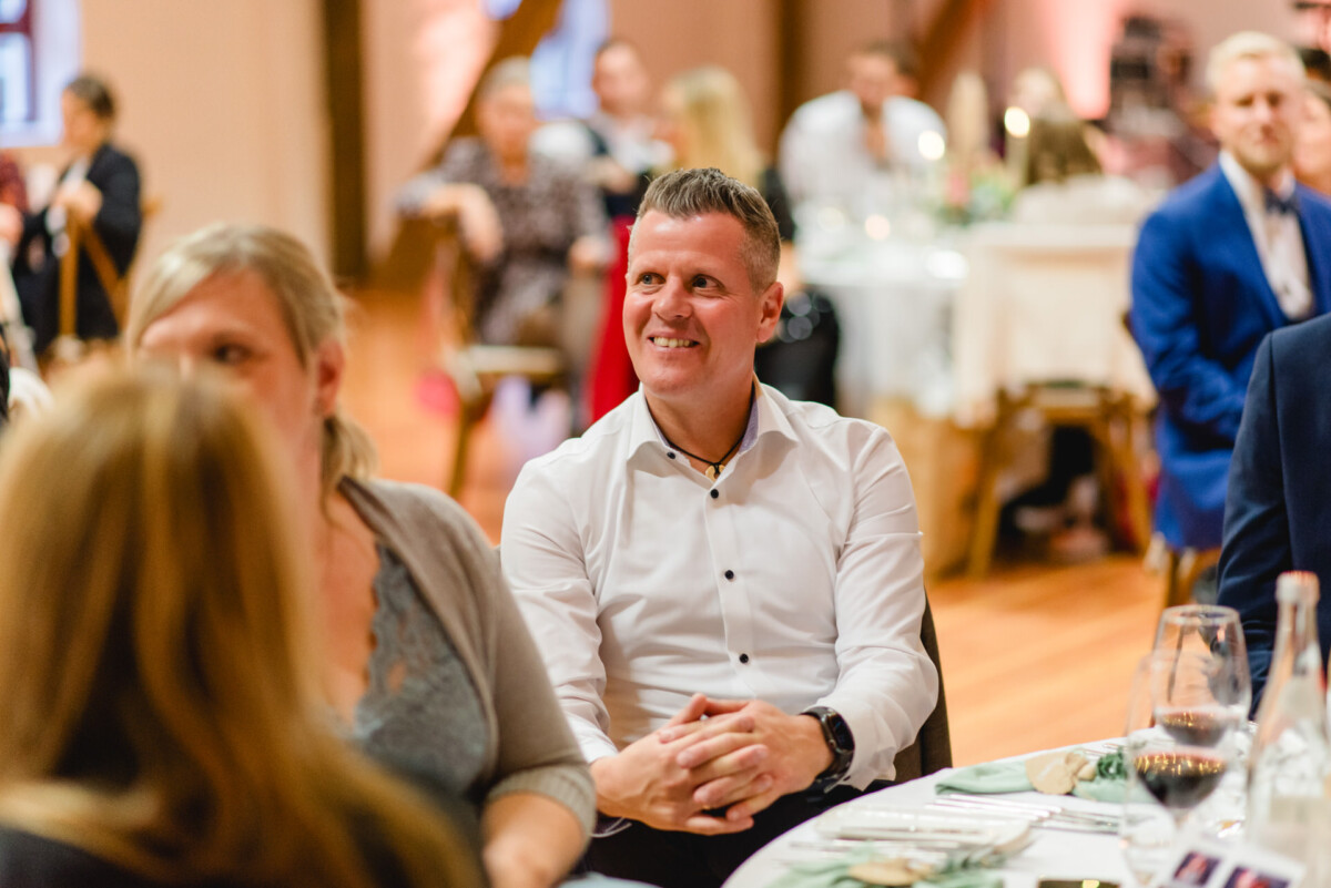 Ein Mann im weißen Hemd sitzt lächelnd bei einer formellen Veranstaltung, umgeben von Menschen, die an Tischen sitzen.