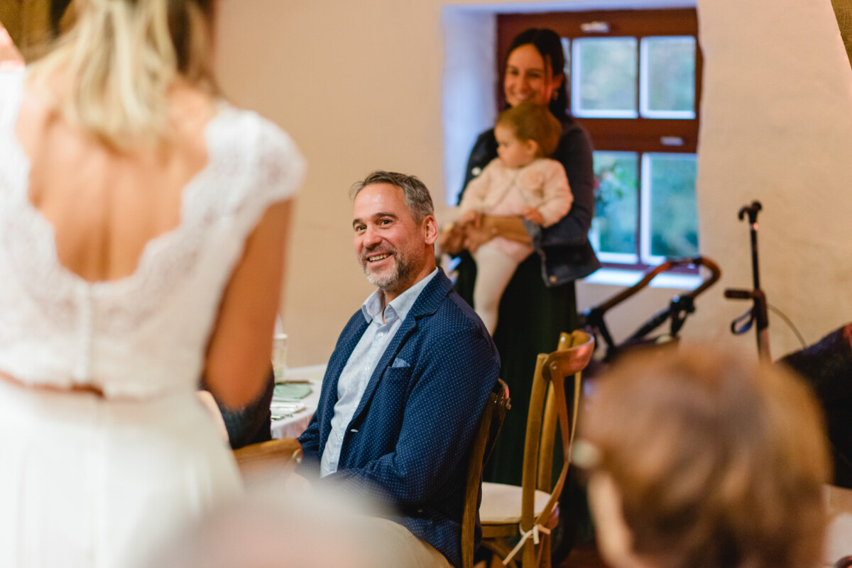 Ein Mann sitzt an einem Tisch und lächelt einer Frau zu, die sich unterhält. Eine andere Frau im Hintergrund hält ein Kind.