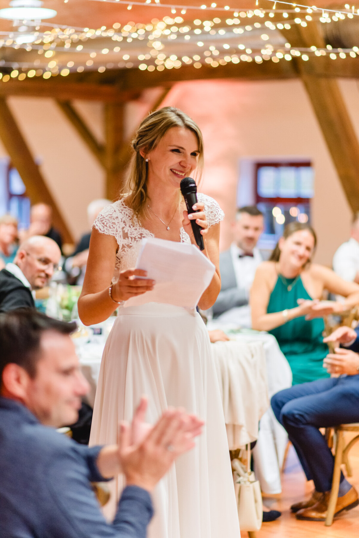 Eine Frau im weißen Kleid hält bei einer Indoor-Veranstaltung mit Mikrofon eine Rede, während die umstehenden Sitzenden applaudieren. Lichterketten schmücken die Holzbalkendecke.