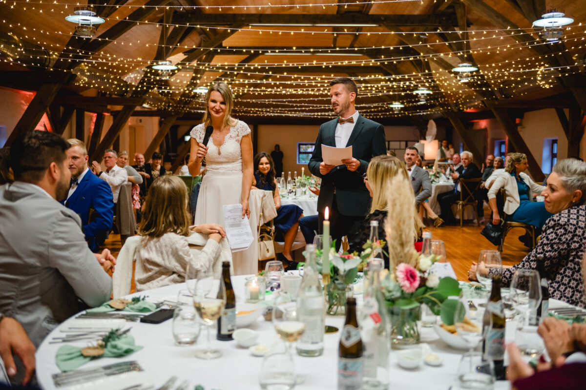 Ein Paar in Abendgarderobe steht und spricht mit Gästen, die an dekorierten Tischen in einem warm beleuchteten Veranstaltungsort sitzen.
