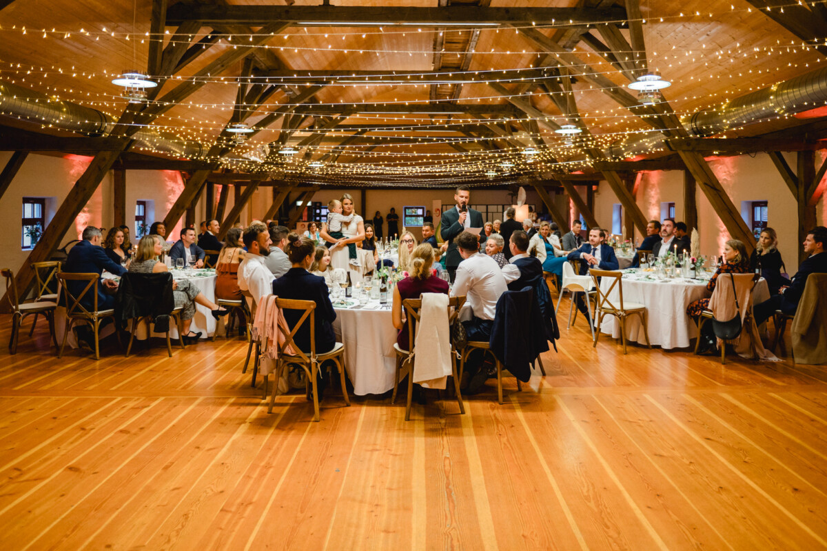 Ein großer Speisesaal mit Gästen an runden Tischen, die mit Lichterketten geschmückt sind. Eine Person steht auf und spricht zu den Gästen, die aufmerksam zuhören.