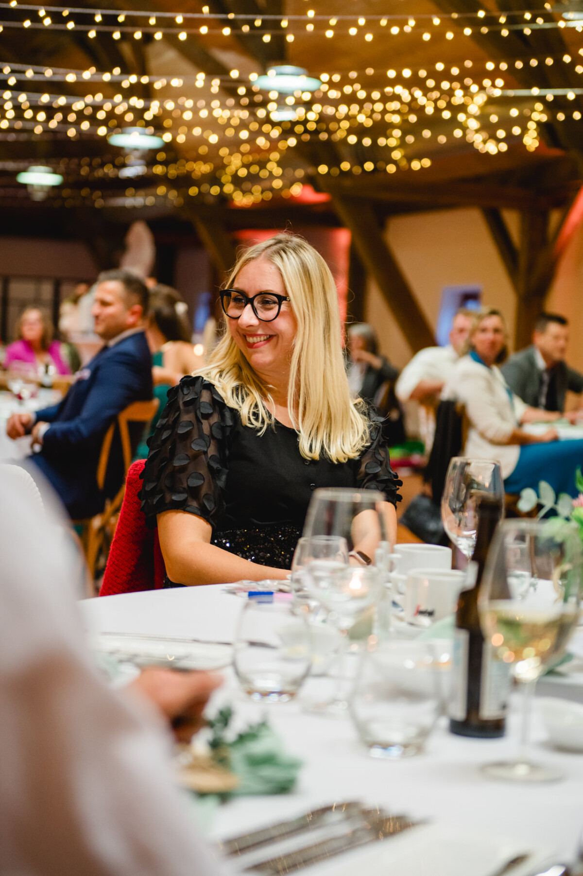 Eine Frau mit Brille sitzt während einer Veranstaltung unter Lichterketten an einem Tisch. Hinter ihr sitzen weitere Gäste an Tischen.