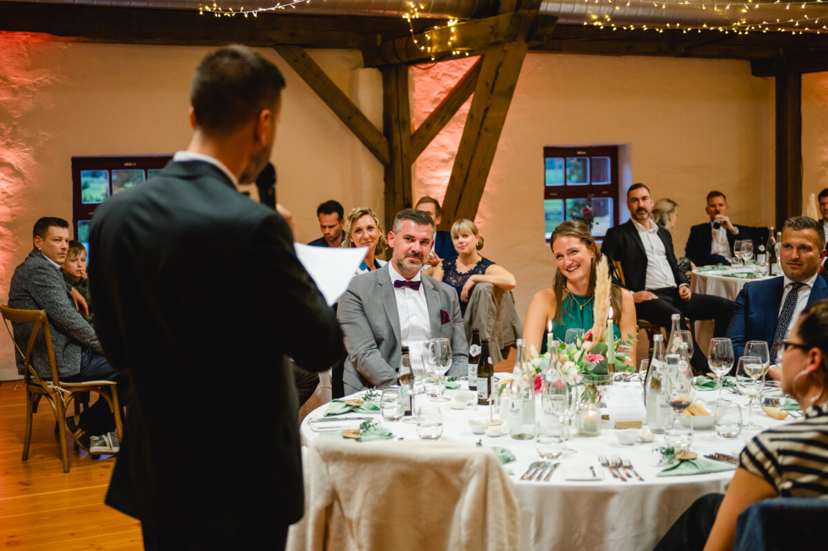 Ein Mann hält bei einer offiziellen Veranstaltung eine Rede, während die Teilnehmer an runden Tischen sitzen, die mit Blumen und Glaswaren dekoriert sind.