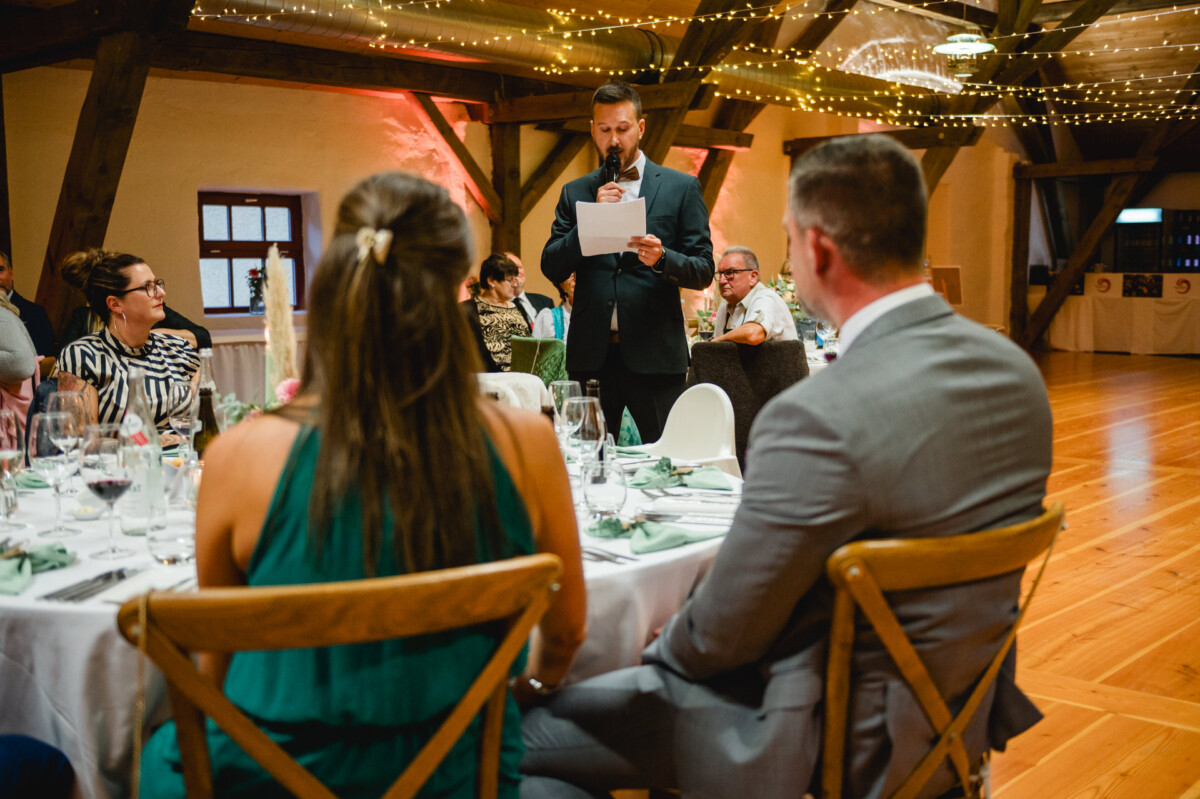 Ein Mann im Anzug spricht bei einer offiziellen Veranstaltung in ein Mikrofon. Die Gäste sitzen an Tischen, die mit Weingläsern und Blumenarrangements dekoriert sind. Von der Decke hängen Lichterketten.