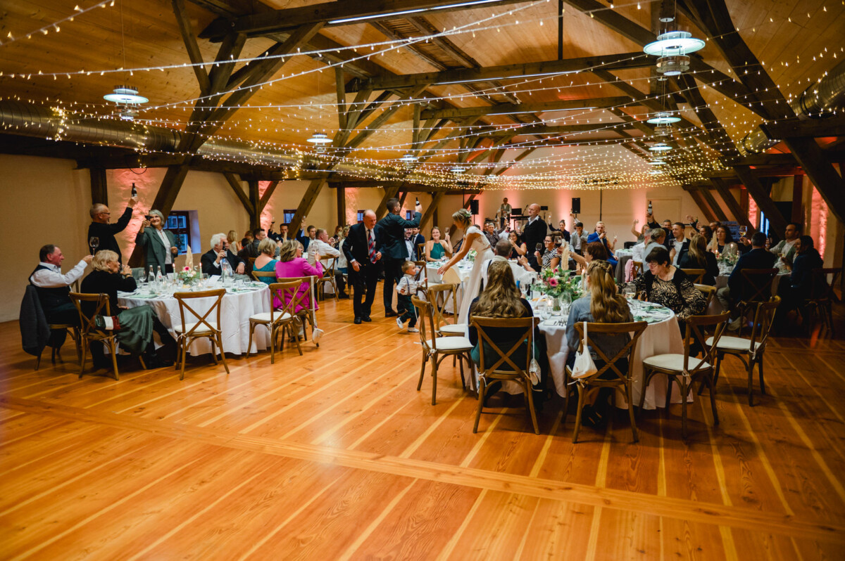 Eine große Gruppe von Menschen sitzt an runden Tischen in einem dekorierten Veranstaltungsort mit Holzboden und Lichterketten. Ein Paar tanzt in der Mitte des Raumes.