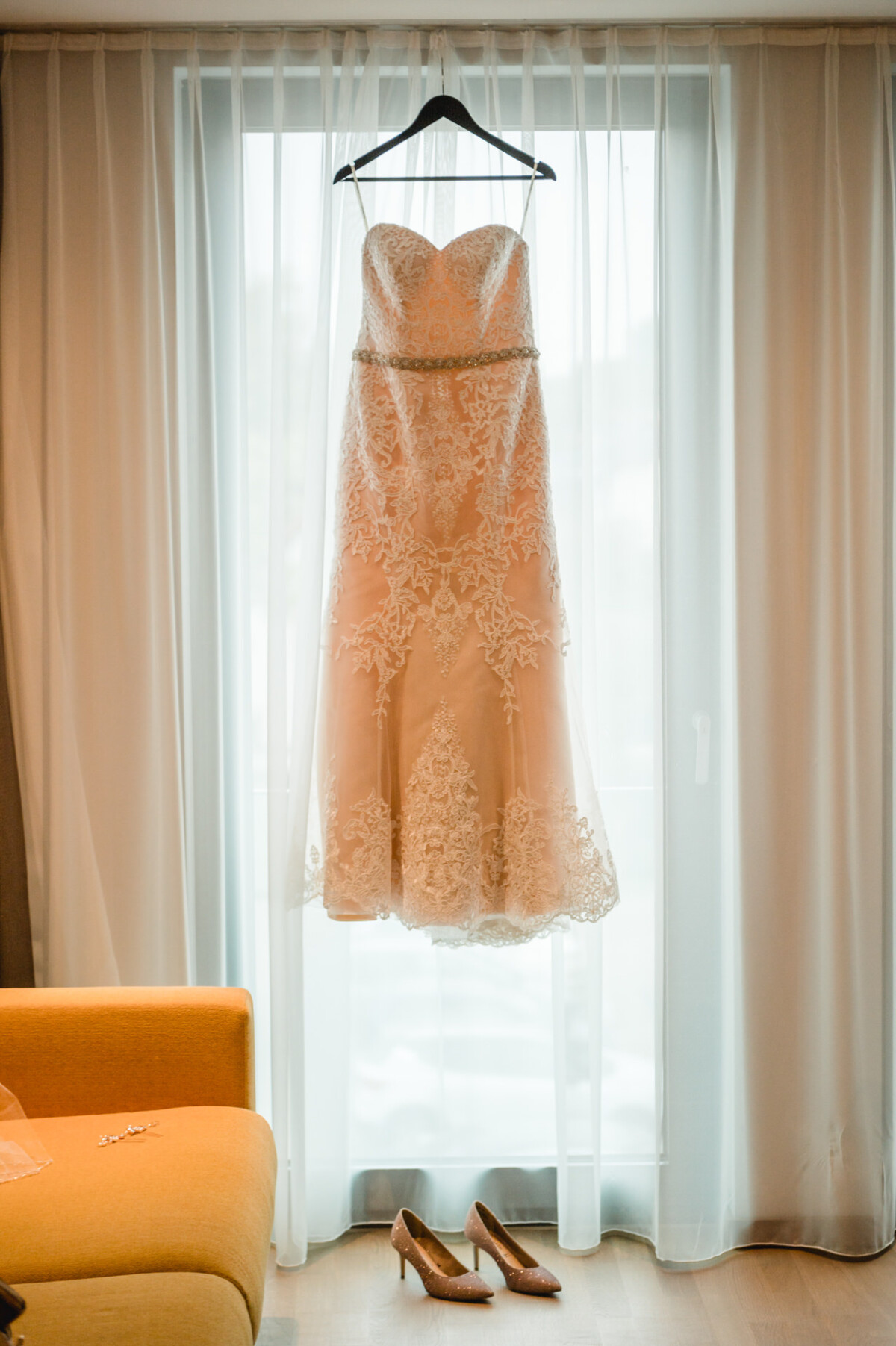 christian-lorenz.net Ein trägerloses Spitzenbrautkleid hängt auf einem Kleiderbügel vor einem Fenster. Darunter stehen ein Paar beige Stöckelschuhe auf dem Boden neben einem orangefarbenen Stuhl.