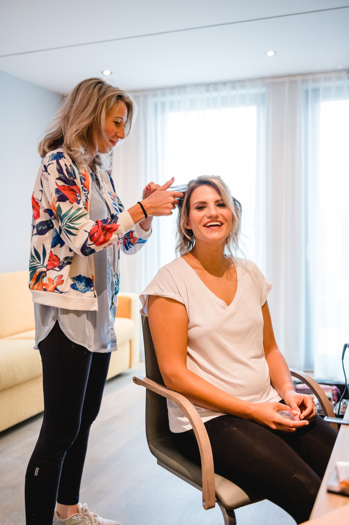 christian-lorenz.net Eine Frau sitzt lächelnd auf einem Stuhl, während eine andere Frau hinter ihr steht und ihr die Haare frisiert. Sie befinden sich in einem gut beleuchteten Raum mit hellen Möbeln.
