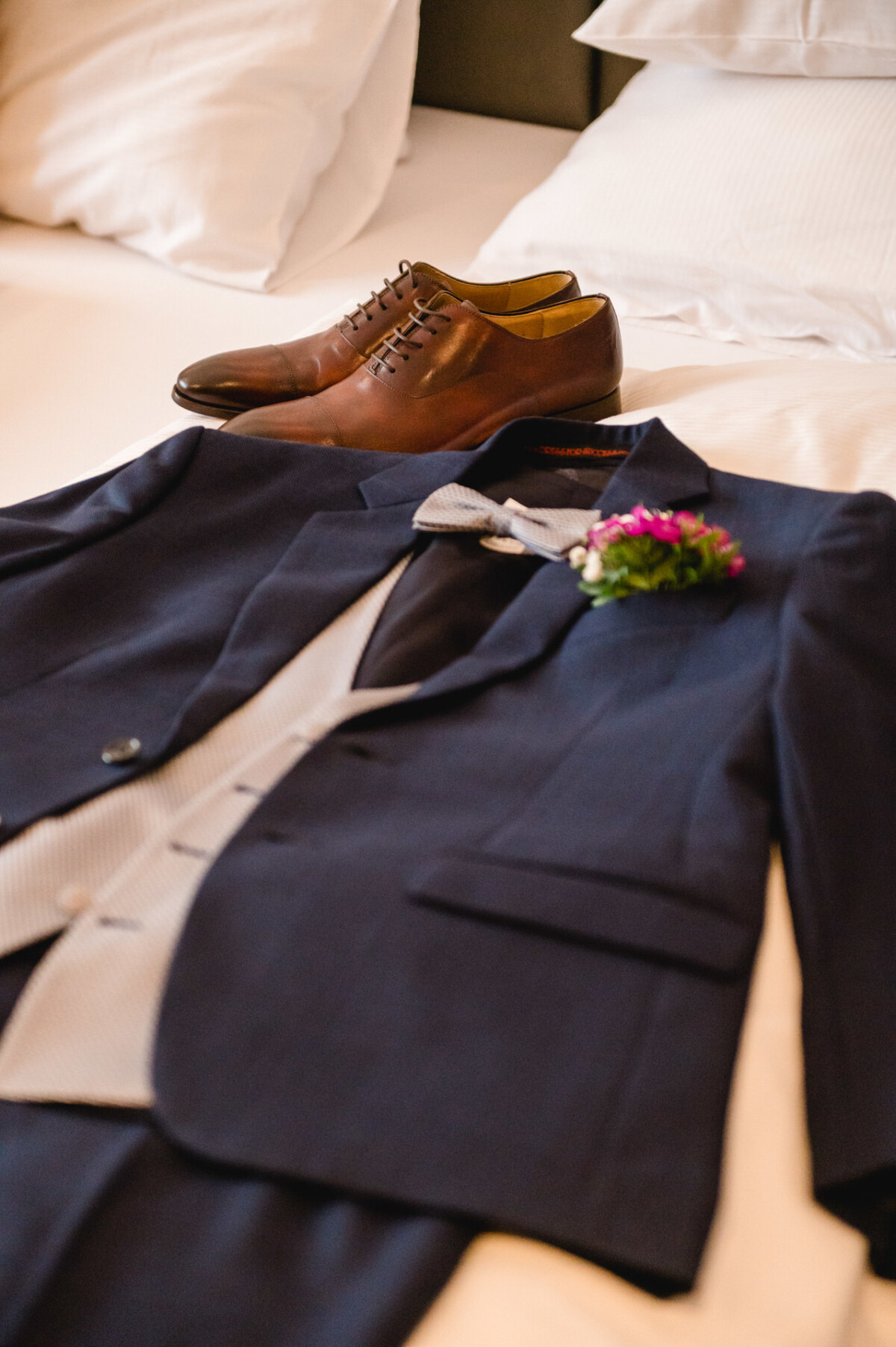 christian-lorenz.net Ein marineblauer Anzug mit grauer Weste, Fliege und Ansteckblume liegt auf einem Bett, im Hintergrund sind braune Lederschuhe zu sehen.